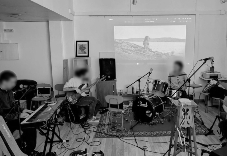 Black and whit photo of three figures with their faces blurred playing music instruments. A bassist, drummer, and a keyboard player with a background of a screen projection