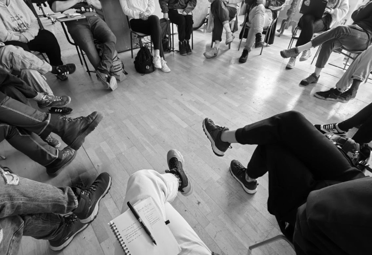Black and white photo of a group of people seated in a circle, only their feet are visible